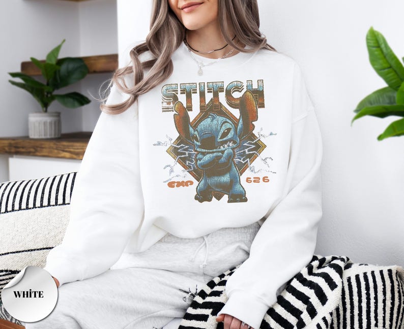 May include: A white crewneck sweatshirt featuring a graphic print of the blue character Stitch with arms crossed. The design includes the text STITCH in a distressed font, a diamond-shaped background, lightning bolts, and the text EXP 626.
