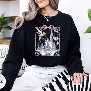 May include: Black sweatshirt featuring a colorful graphic of a fairytale castle, fireworks, and a fairy. The design includes a pink bow and floral accents. The sweatshirt is a solid black color.