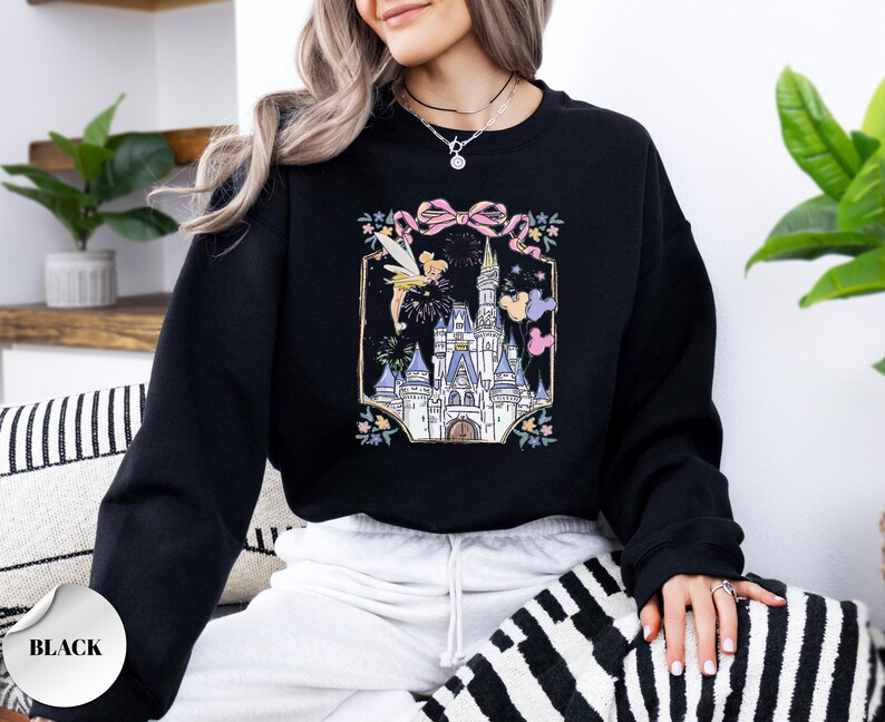 May include: Black sweatshirt featuring a colorful graphic of a fairytale castle, fireworks, and a fairy. The design includes a pink bow and floral accents. The sweatshirt is a solid black color.