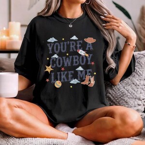 May include: A black short-sleeved t-shirt featuring the text "YOU'RE A COWBOY LIKE ME" in a dark blue font. The design includes graphic illustrations of a brown cowboy hat, a rocket, cowboy boots, a ball, stars, and light blue clouds.