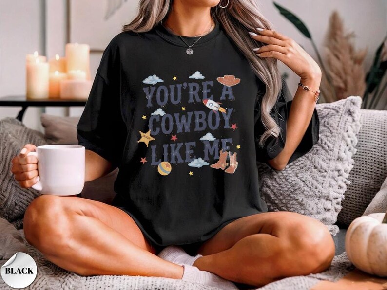 May include: A black short-sleeved t-shirt featuring the text "YOU'RE A COWBOY LIKE ME" in a dark blue font. The design includes graphic illustrations of a brown cowboy hat, a rocket, cowboy boots, a ball, stars, and light blue clouds.