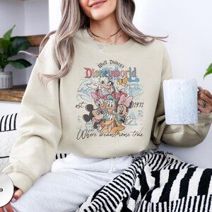 Op de afbeelding: Zandkleurige sweatshirt met een retro afbeelding van Disney-personages, waaronder Mickey Mouse, Minnie Mouse en Donald Duck. De afbeelding bevat de tekst "Walt Disney's Disneyworld est. 1971" en "Where dreams come true."