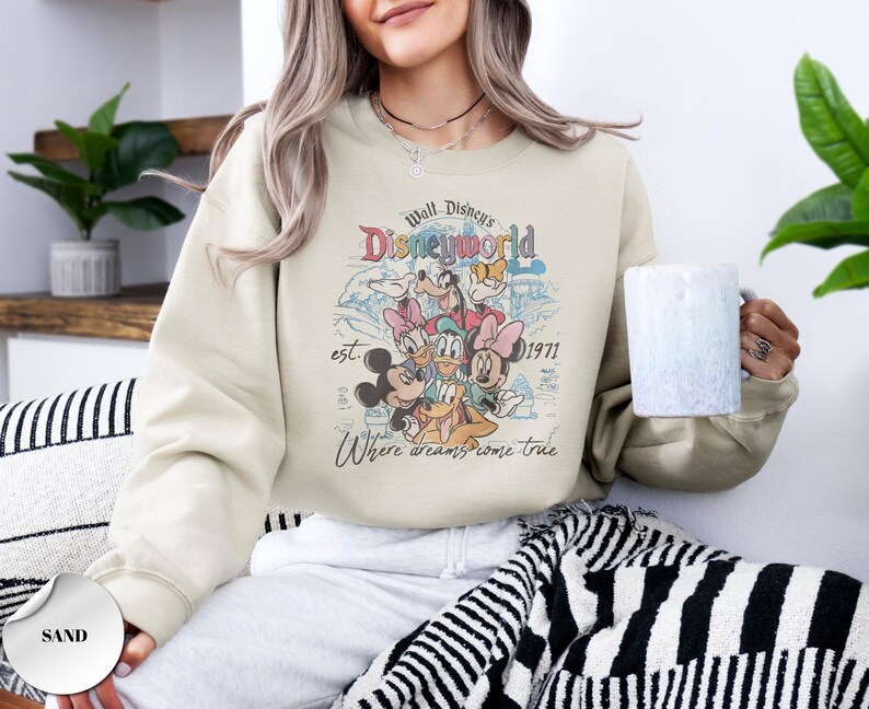 Op de afbeelding: Zandkleurige sweatshirt met een retro afbeelding van Disney-personages, waaronder Mickey Mouse, Minnie Mouse en Donald Duck. De afbeelding bevat de tekst "Walt Disney's Disneyworld est. 1971" en "Where dreams come true."