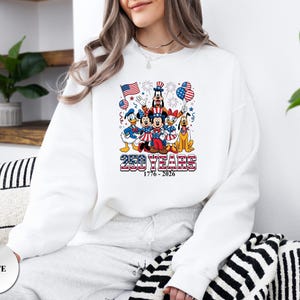 May include: A white long-sleeve sweatshirt featuring a graphic of Disney characters in patriotic attire with American flags, balloons, and fireworks. The text reads 250 YEARS 1776 - 2026 in a red, white, and blue striped font style.