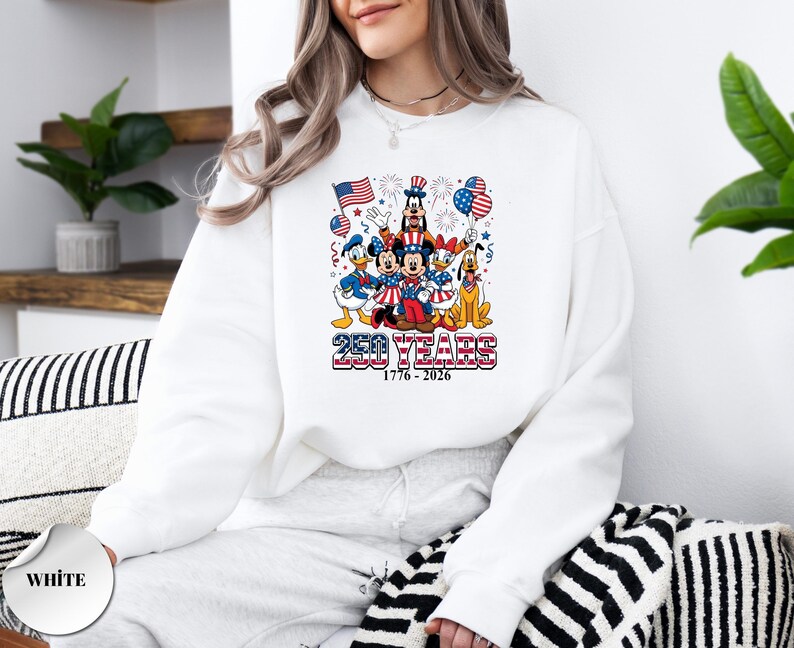 May include: A white long-sleeve sweatshirt featuring a graphic of Disney characters in patriotic attire with American flags, balloons, and fireworks. The text reads 250 YEARS 1776 - 2026 in a red, white, and blue striped font style.