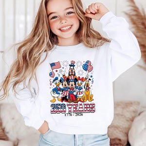 May include: A white long-sleeve sweatshirt featuring a graphic of Disney characters including Mickey Mouse, Minnie Mouse, Donald Duck, Daisy Duck, Goofy, and Pluto dressed in patriotic attire with American flags, balloons, and the text 250 YEARS 1776-2026.