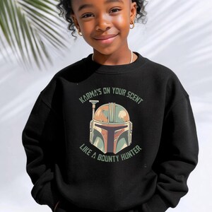 Puede incluir: Una sudadera negra de manga larga y cuello redondo con un estampado gr&aacute;fico de un casco estilizado en tonos verde, beige y marr&oacute;n. El texto "KARMA'S ON YOUR SCENT" aparece arqueado sobre el casco y "LIKE A BOUNTY HUNTER" debajo del mismo.