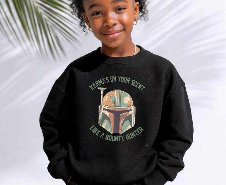 Puede incluir: Una sudadera negra de manga larga y cuello redondo con un estampado gr&aacute;fico de un casco estilizado en tonos verde, beige y marr&oacute;n. El texto "KARMA'S ON YOUR SCENT" aparece arqueado sobre el casco y "LIKE A BOUNTY HUNTER" debajo del mismo.