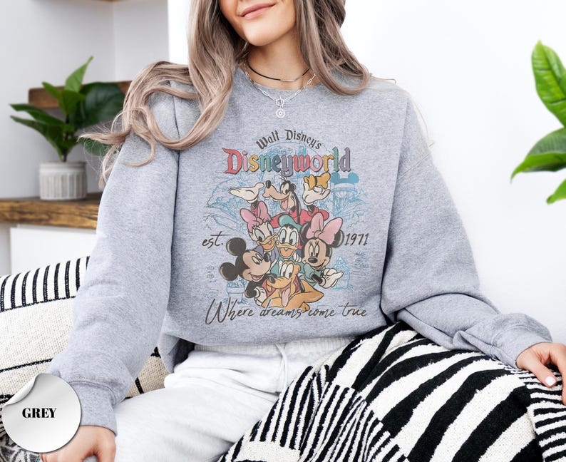 Op de afbeelding: Een grijze sweatshirt met een kleurrijke afbeelding van Disney-personages, waaronder Mickey Mouse, Minnie Mouse en Donald Duck. De afbeelding luidt "Walt Disney's Disneyworld est. 1971" en "Where dreams come true."