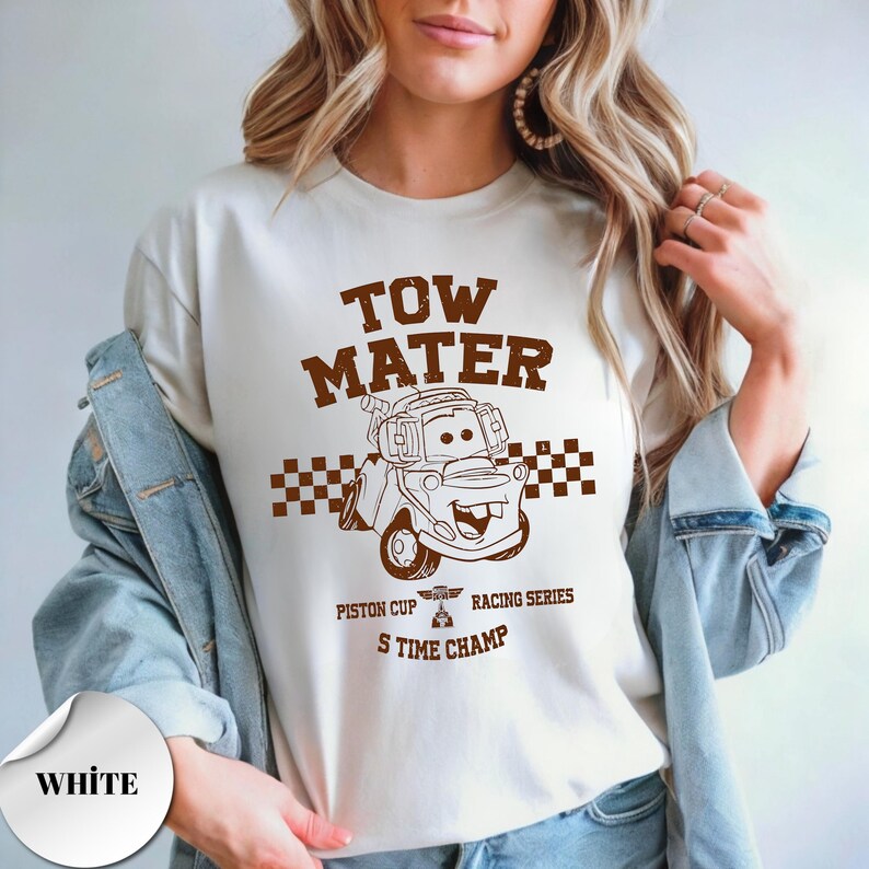 May include: A white t-shirt featuring a brown graphic of the character Tow Mater from the movie Cars. The design includes the text TOW MATER, a checkered flag pattern, and the words PISTON CUP RACING SERIES S TIME CHAMP in brown ink.