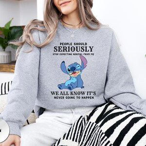 May include: A grey crewneck sweatshirt featuring a graphic of the blue character Stitch from Lilo and Stitch. The text reads: PEOPLE SHOULD SERIOUSLY STOP EXPECTING NORMAL FROM ME WE ALL KNOW IT'S NEVER GOING TO HAPPEN in black lettering.
