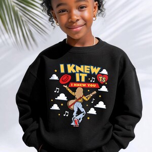 May include: A black crewneck sweatshirt featuring a graphic print with the text I KNEW IT and I KNEW YOU. The design includes a person with long hair playing a guitar, a red hat, a heart with the letters TS, white clouds, and musical notes.