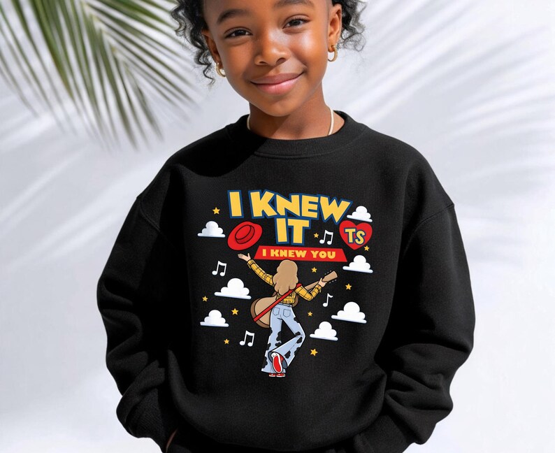 May include: A black crewneck sweatshirt featuring a graphic print with the text I KNEW IT and I KNEW YOU. The design includes a person with long hair playing a guitar, a red hat, a heart with the letters TS, white clouds, and musical notes.