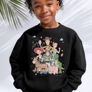 May include: A black crewneck sweatshirt featuring a colorful graphic print of Toy Story characters including Woody, Buzz Lightyear, Jessie, Rex, Hamm, Mr. Potato Head, Bo Peep, and Forky, surrounded by small stars, clouds, and character names.