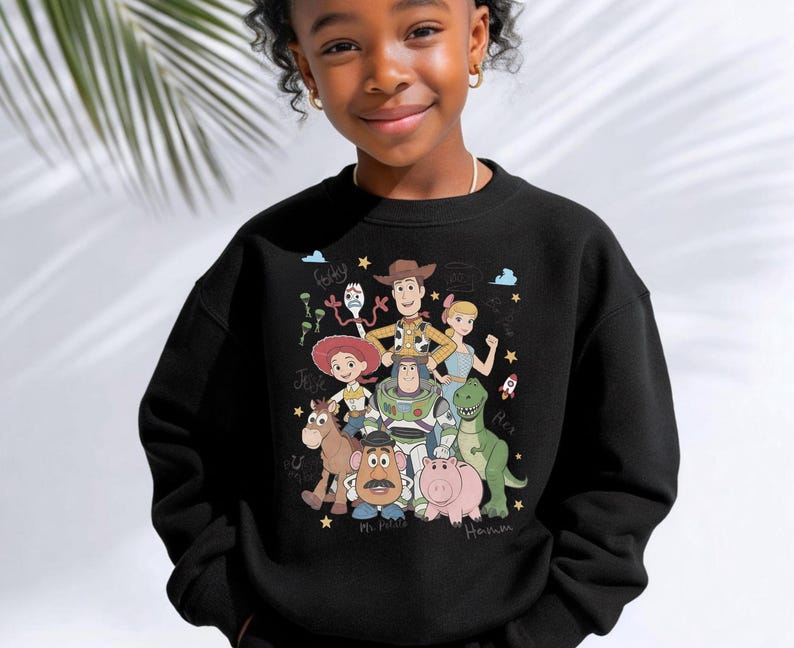 May include: A black crewneck sweatshirt featuring a colorful graphic print of Toy Story characters including Woody, Buzz Lightyear, Jessie, Rex, Hamm, Mr. Potato Head, Bo Peep, and Forky, surrounded by small stars, clouds, and character names.