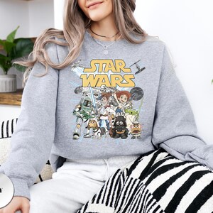 Puede incluir: Una sudadera gris de cuello redondo con un estampado gr&aacute;fico de personajes de Star Wars reimaginados como figuras de Toy Story. El dise&ntilde;o incluye el logotipo amarillo de Star Wars sobre ilustraciones de personajes como Jedi, Sith y alien&iacute;genas.
