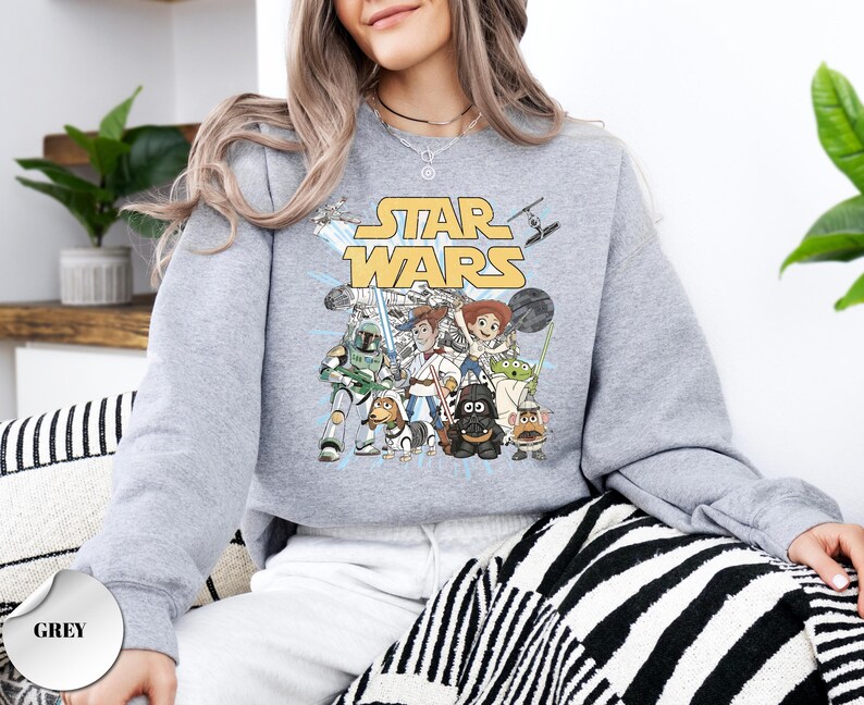Puede incluir: Una sudadera gris de cuello redondo con un estampado gr&aacute;fico de personajes de Star Wars reimaginados como figuras de Toy Story. El dise&ntilde;o incluye el logotipo amarillo de Star Wars sobre ilustraciones de personajes como Jedi, Sith y alien&iacute;genas.