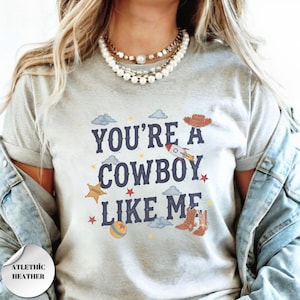 May include: A light gray t-shirt featuring the dark blue text "YOU'RE A COWBOY LIKE ME" surrounded by colorful illustrations of a brown cowboy hat, a rocket, a yellow sheriff star, a ball, and small stars and clouds scattered across the front.