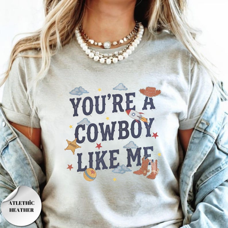 May include: A light gray t-shirt featuring the dark blue text "YOU'RE A COWBOY LIKE ME" surrounded by colorful illustrations of a brown cowboy hat, a rocket, a yellow sheriff star, a ball, and small stars and clouds scattered across the front.