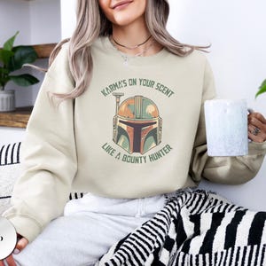 Puede incluir: Una sudadera de cuello redondo color arena con un gr&aacute;fico de un casco de cazarrecompensas en tonos verde, naranja y marr&oacute;n. El texto "KARMA'S ON YOUR SCENT LIKE A BOUNTY HUNTER" est&aacute; impreso en letras may&uacute;sculas verdes en arco sobre el casco.