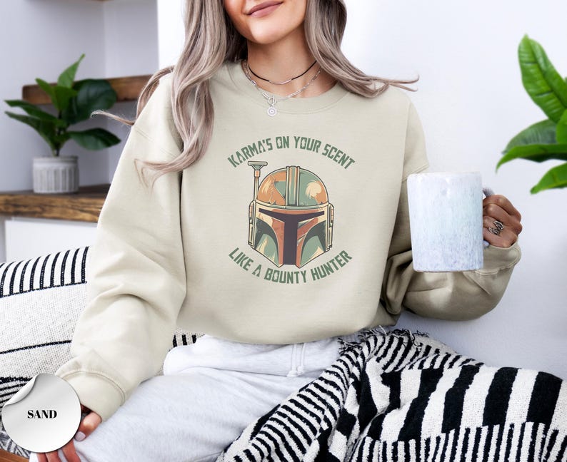 Puede incluir: Una sudadera de cuello redondo color arena con un gr&aacute;fico de un casco de cazarrecompensas en tonos verde, naranja y marr&oacute;n. El texto "KARMA'S ON YOUR SCENT LIKE A BOUNTY HUNTER" est&aacute; impreso en letras may&uacute;sculas verdes en arco sobre el casco.