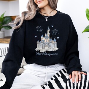 May include: A black long-sleeve sweatshirt featuring a graphic print of a light blue and white castle with fireworks and the text Walt Disney World. The design includes small blue Mickey Mouse head silhouettes surrounding the castle illustration.