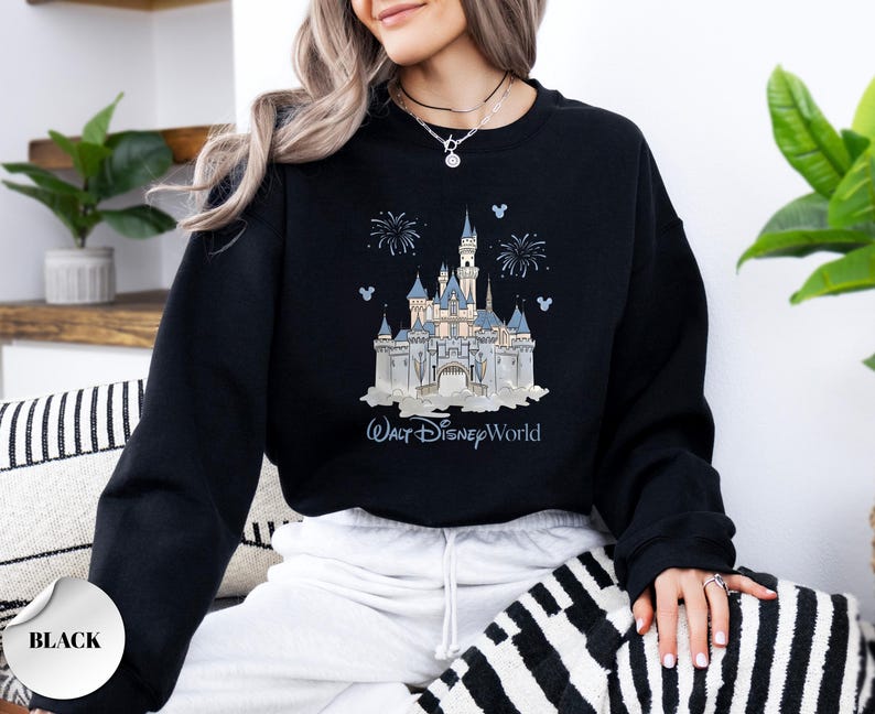 May include: A black long-sleeve sweatshirt featuring a graphic print of a light blue and white castle with fireworks and the text Walt Disney World. The design includes small blue Mickey Mouse head silhouettes surrounding the castle illustration.