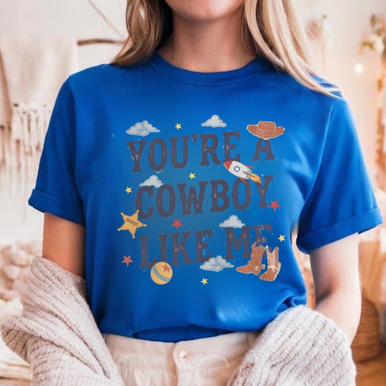 May include: A bright blue short-sleeved t-shirt featuring the text "YOU'RE A COWBOY LIKE ME" in dark letters. The design includes illustrations of a brown cowboy hat, a rocket ship, a yellow sheriff star, a ball, and brown cowboy boots.