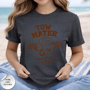 May include: A dark grey heather t-shirt featuring a brown graphic of the character Tow Mater from the movie Cars. The design includes the text TOW MATER, a checkered flag pattern, and the words PISTON CUP RACING SERIES 5 TIME CHAMP.