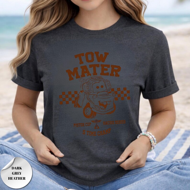 May include: A dark grey heather t-shirt featuring a brown graphic of the character Tow Mater from the movie Cars. The design includes the text TOW MATER, a checkered flag pattern, and the words PISTON CUP RACING SERIES 5 TIME CHAMP.