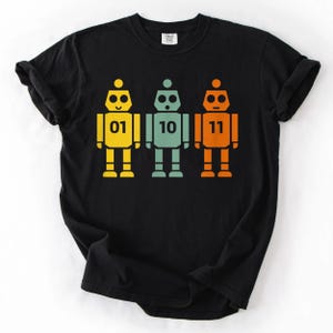 May include: Black t-shirt featuring three colorful robot illustrations. Each robot is a different color: yellow, teal, and orange. The robots have the numbers 01, 10, and 11 printed on their chests. The design is simple and graphic.
