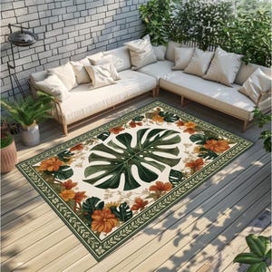 Könnte beinhalten: Rechteckiger Outdoor-Teppich mit einem großen grünen Monstera-Blatt-Design in der Mitte, umgeben von orangefarbenen Blumen und grünen Blättern auf cremefarbenem Hintergrund. Der Teppich hat einen grünen Rand mit einem Blättermuster. Er liegt auf einer Holzterrasse.