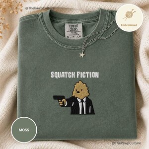 May include: A moss green crewneck t-shirt featuring an embroidered graphic of a tan creature in a black suit holding a black handgun. Above the figure, the text reads SQUATCH FICTION in white. The shirt has a Comfort Colors brand label.