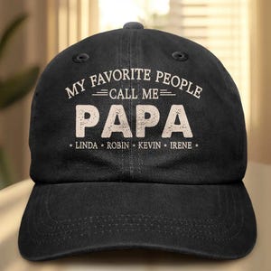 May include: A black baseball cap featuring white text that reads MY FAVORITE PEOPLE CALL ME PAPA, with the names LINDA, ROBIN, KEVIN, and IRENE printed in a smaller font below. The cap has a structured crown, a curved brim, and eyelet vents.
