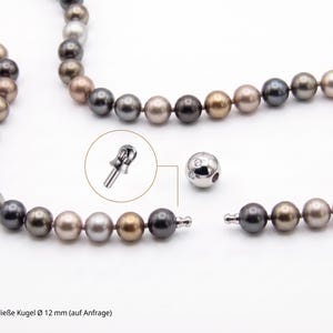 Puede incluir: Un collar de perlas multicolores con un cierre plateado. Las perlas son en tonos grises, marrones y dorados. El cierre es una bola plateada con pequeñas piedras transparentes. El collar se muestra desabrochado, con un primer plano del mecanismo de cierre.