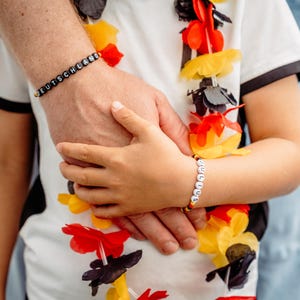 Könnte beinhalten: Ein weißes T-Shirt mit schwarzem Saum an den Ärmeln, getragen mit einer Blumenkette aus schwarzen, roten und gelben Blütenblättern. Zwei Perlenarmbänder sind sichtbar: eines mit schwarzen Perlen, die DEUTSCHLAND buchstabieren, und eines mit weißen.