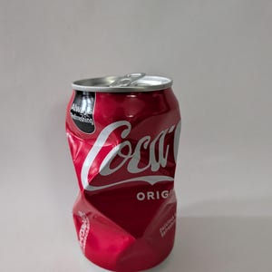 Può includere: Una lattina di Coca-Cola rossa schiacciata con il marchio in bianco. La lattina è ammaccata e sgualcita, con le parole "Always Refreshing" e "Original" visibili. Su sfondo bianco.