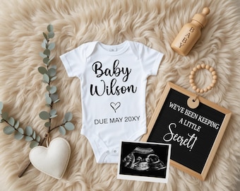 May include: White onesie with the text "Baby Wilson" and a heart, with the due date "DUE MAY 20XY". A black chalkboard sign reads "We've been keeping a little Secret!" with a wooden baby bottle and a wooden bead necklace.