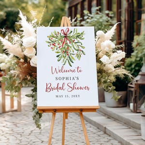 May include: A white sign with a watercolor illustration of mistletoe with a red ribbon. The sign reads "Welcome to Sophia's Bridal Shower May 15, 20XY". The sign is on an easel in front of a brick building.
