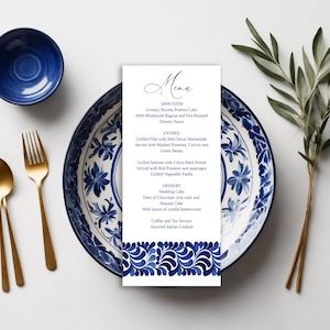 May include: A wedding menu card with a blue and white floral design, placed on a matching plate. Gold utensils and a small blue bowl are arranged nearby. A sprig of greenery adds a natural touch to the elegant table setting.