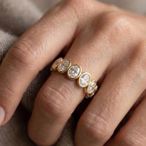 Peut inclure: Une bague en or ornée d'une série de diamants de taille ovale sertis dans un style de lunette. Les diamants sont clairs et étincelants, disposés en une rangée continue autour de l'anneau. La bague est portée sur une main.