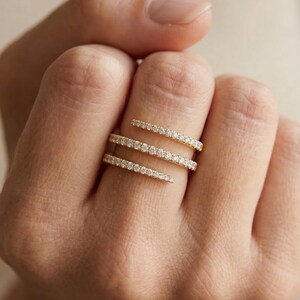 Könnte beinhalten: Drei goldfarbene Ringe, jeder mit einer Reihe kleiner, funkelnder Steine besetzt, werden auf einem Finger gezeigt. Die Ringe sind in einem spiralförmigen Design angeordnet, wodurch ein geschichteter Effekt entsteht. Der Hintergrund ist ein weicher, neutraler Ton.