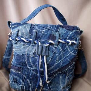 May include: A patchwork denim backpack with a top handle and adjustable shoulder straps. The bag features a drawstring closure with white and blue rope detailing. The denim is a mix of light and dark blue shades, with white stitching.