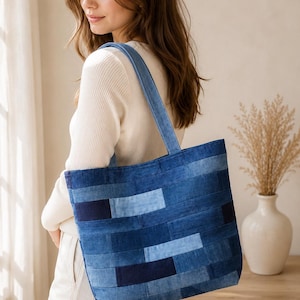 May include: A rectangular tote bag constructed from horizontal strips of denim fabric in varying shades of blue, ranging from light sky blue to deep navy. The bag features two matching denim shoulder straps and a structured, flat-bottomed design.