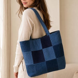 Puede incluir: Un bolso tote confeccionado en tela de mezclilla con un diseño de patchwork de paneles cuadrados alternados en azul claro y azul oscuro. El bolso incluye dos correas para el hombro y una forma rectangular, mostrando una construcción textil clásica.