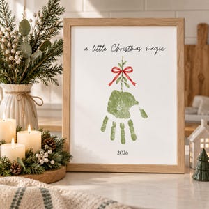 Może przedstawiać: Oprawiony wydruk przedstawiający zielony odcisk dłoni w kształcie drzewa z czerwoną kokardą na górze. Tekst brzmi a little Christmas magic na górze i 2026 na dole. Wydruk znajduje się w jasnej drewnianej ramie na białej powierzchni.