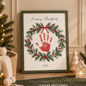 May include: A framed print featuring a watercolor Christmas wreath with green pine needles and red berries. A red handprint is centered within the wreath. The text Merry Christmas is at the top and 2026 is at the bottom. The text 2 SIZES INCLUDED A4 US Letter PRINT ADD A HANDPRINT DISPLAY is at the bottom.