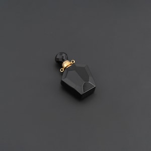 May include: A small, faceted black perfume bottle pendant with a geometric shape. The bottle features a gold-toned metal neck and a matching black faceted stopper, with two small gold-toned loops attached to the neck for hanging as jewelry.