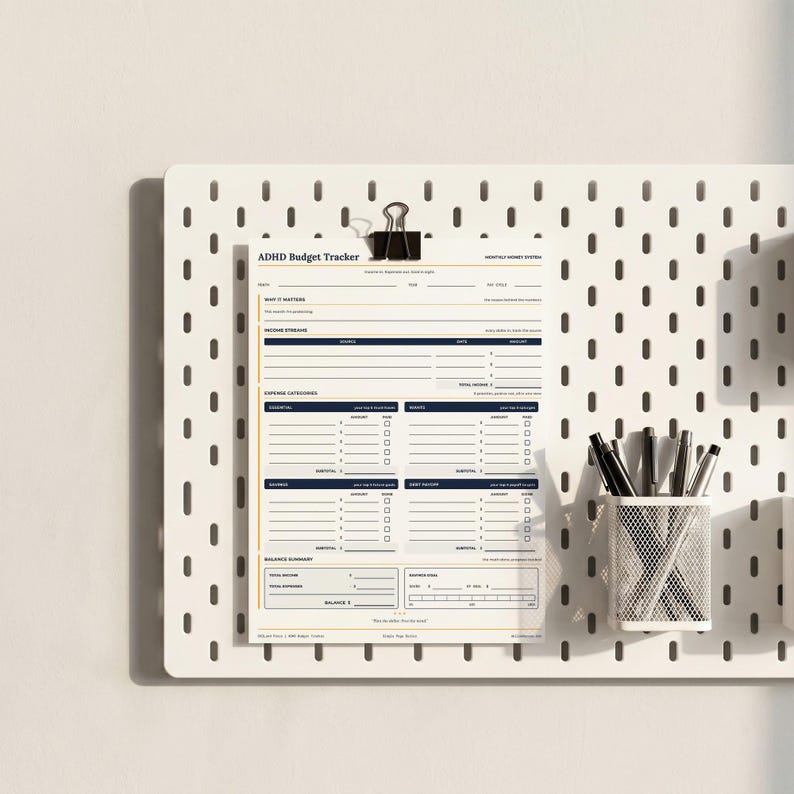May include: A printed ADHD Budget Tracker sheet with sections for income streams, expense categories, and a balance summary, secured by a black binder clip to a white pegboard, accompanied by a white mesh pen holder containing several pens.