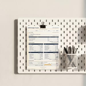 May include: A white ADHD Budget Tracker paper form with dark blue and gold text, held by a black binder clip on a white pegboard. The form includes sections for income streams, expense categories, and a balance summary with printed financial data.